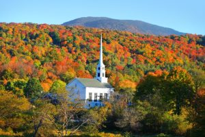 Witte houten kerk met hoge torenspits omringd door bomen in feloranje en rode herfstkleuren tegen een berg in Vermont