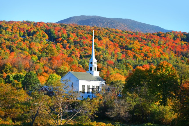 Witte houten kerk met hoge torenspits omringd door bomen in feloranje en rode herfstkleuren tegen een berg in Vermont