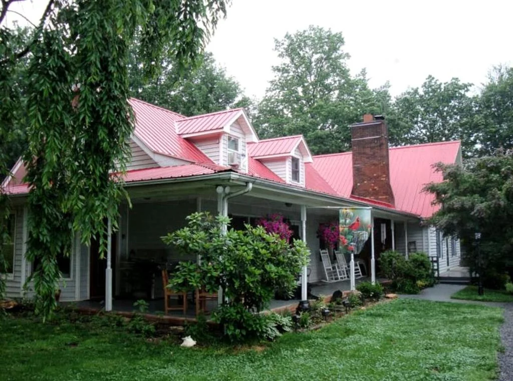 Wit houten huis met een rood metalen dak, schoorsteen en een overdekte veranda omgeven door groen in Virginia