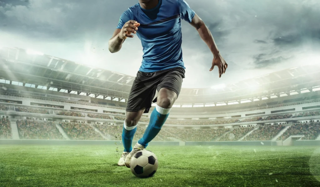 Voetballer in blauw shirt raakt de bal met zijn voet in een fel verlicht stadion met vage tribunes op de achtergrond
