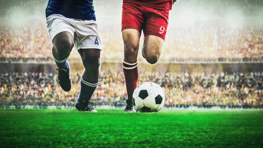 Twee spelers in verschillende shirts strekken hun benen naar de bal tijdens een duel op het speelveld