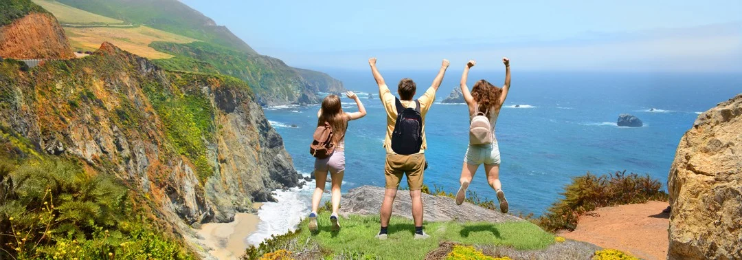 Drie reizigers met rugzakken juichen boven op een klif met uitzicht over de oceaan langs de kust van Californië