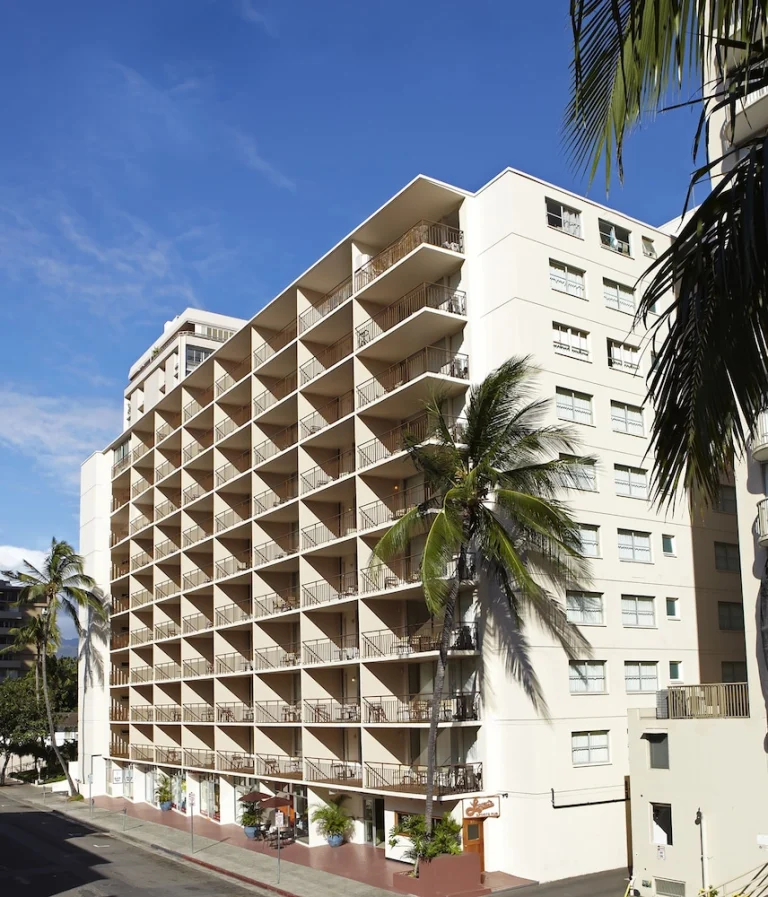 Hoog wit hotelgebouw met rijen balkons en een palmboom op de voorgrond onder een blauwe lucht in Waikiki