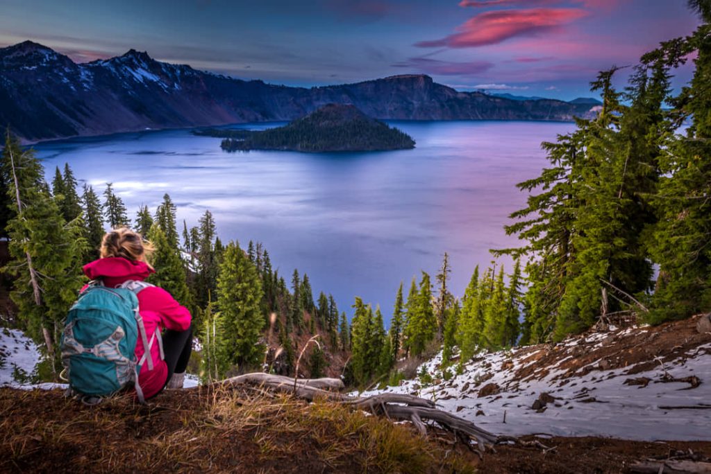 Een wandelaar met rugzak zit op een rotsrichel met uitzicht over het diepblauwe water van Crater Lake en Wizard Island