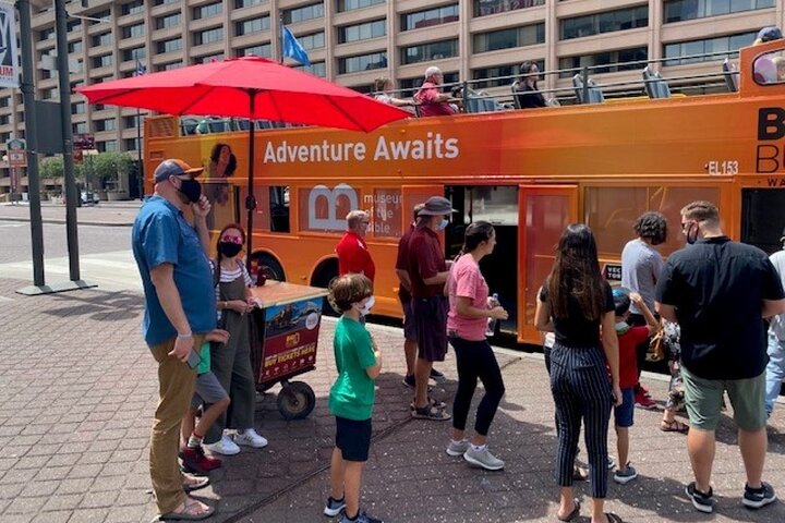 Rij bezoekers met mondkapjes wacht onder een rode parasol bij een oranje dubbeldekker sightseeingbus in de stad
