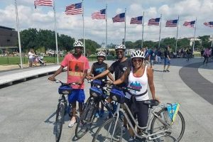 Vier fietsers met helmen poseren naast hun fietsen voor een rij wapperende Amerikaanse vlaggen op de National Mall