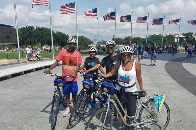 Vier fietsers met helmen poseren naast hun fietsen voor een rij wapperende Amerikaanse vlaggen op de National Mall