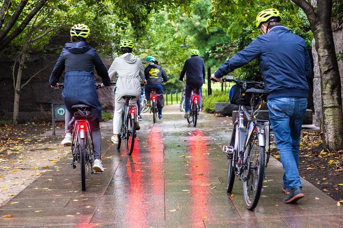 Groep fietsers met helmen en rode achterlichten rijdt over een nat pad tussen bomen in een park