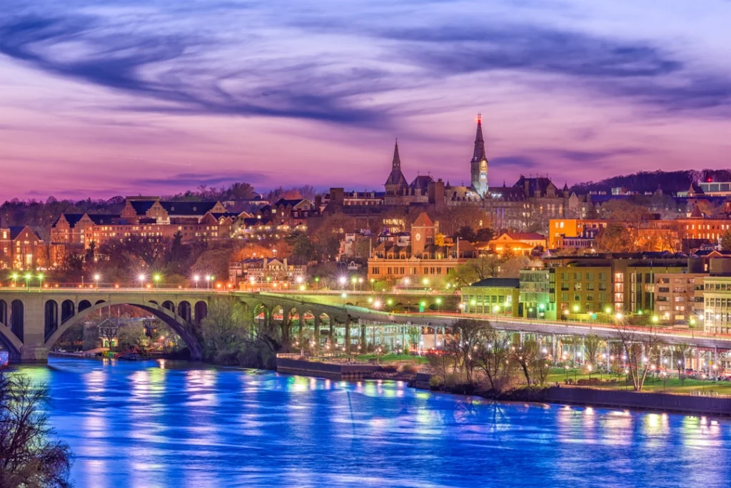 Verlichte gebouwen en kerktorens van Georgetown weerspiegeld in de rivier, met een verlichte brug op de voorgrond bij schemering