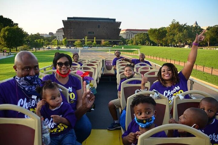 Vrolijk gezin in paarse shirts zit op een open sightseeingbus met op de achtergrond het bronskleurige gebouw van het National Museum of African American History