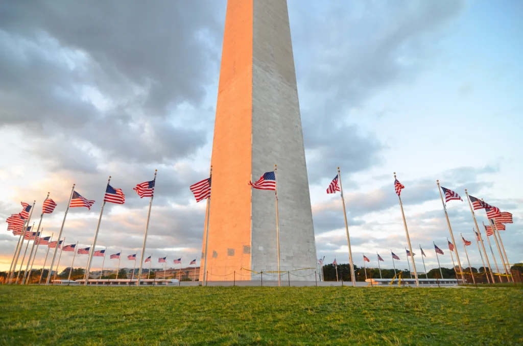 Het Washington Monument in Washington D.C. gehuld in warm ochtendlicht, omringd door een cirkel van wapperende Amerikaanse vlaggen op een grasveld