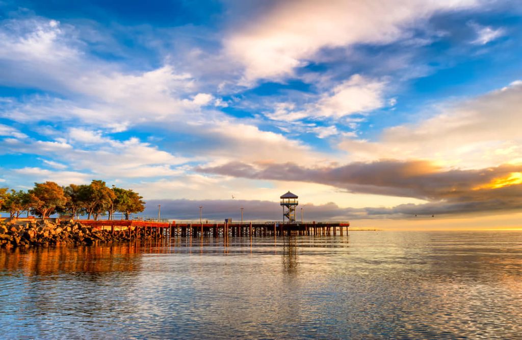 Lange houten pier met een kleine uitkijktoren die uitsteekt in kalm water onder een bewolkte lucht bij zonsondergang in Washington
