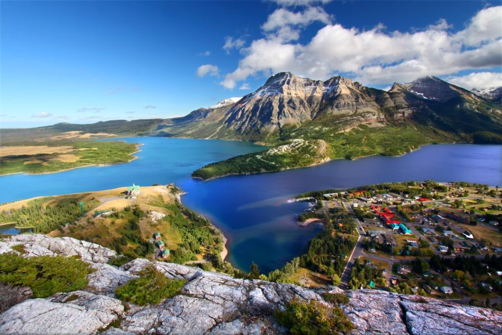 Luchtfoto vanaf een rotsachtige uitkijkpost over het dorp Waterton met een blauw meer en steile bergen eromheen