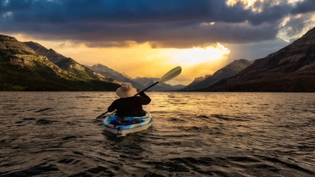 Een persoon met hoed peddelt in een kajak over het rimpelende water van Waterton Lake, omringd door bergen bij zonsondergang