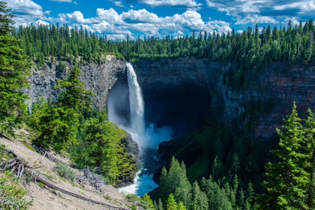 Hoge waterval stort van een rotsrand naar beneden in een brede beboste canyon onder een blauwe hemel met wolken