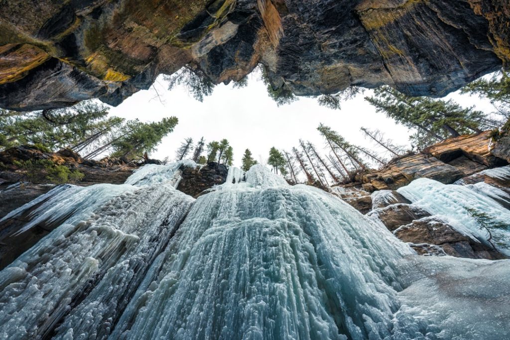 Onderaanzicht van een bevroren waterval met grote ijspegels tussen rotswanden en dennenbomen tegen een lichte lucht