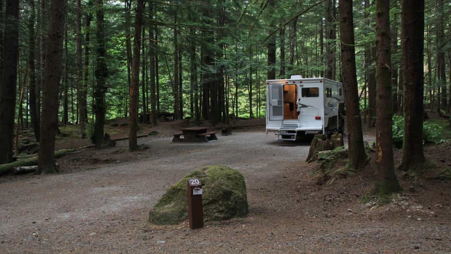 Witte camper staat geparkeerd op een bosrijke camping met een picknicktafel en een grote mosbedekte steen op de voorgrond