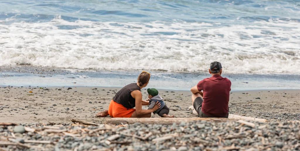 Ouders en een kind zitten met de rug naar de camera op een kiezelstrand terwijl golven aanrollen