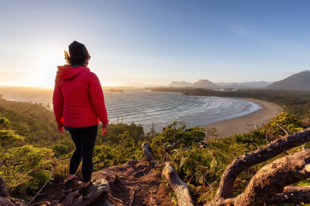 Vrouw in rode jas staat op een rotsachtige uitkijkpost en kijkt uit over een golvende strandbaai bij zonsopgang
