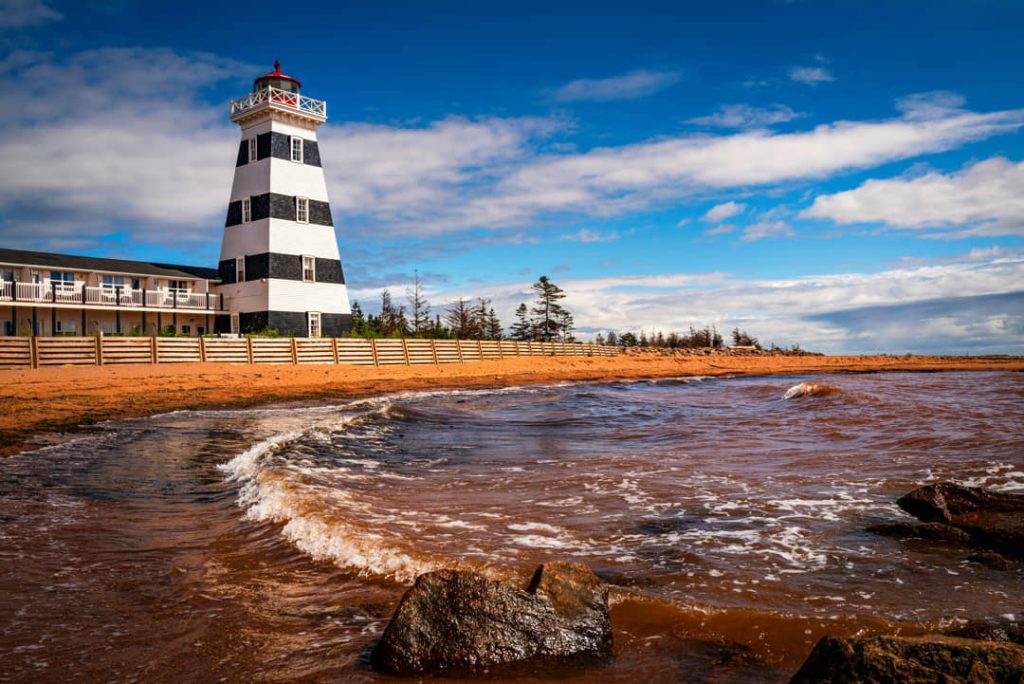 Zwartwit gestreepte vuurtoren naast een hotel aan het strand van West Point op Prince Edward Island bij golvend water