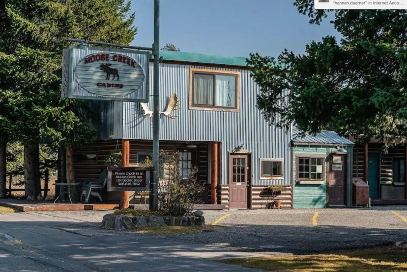 Gevel van een houten en metalen gebouw met het uithangbord Moose Creek Cabins en een parkeerplaats ervoor onder dennenbomen