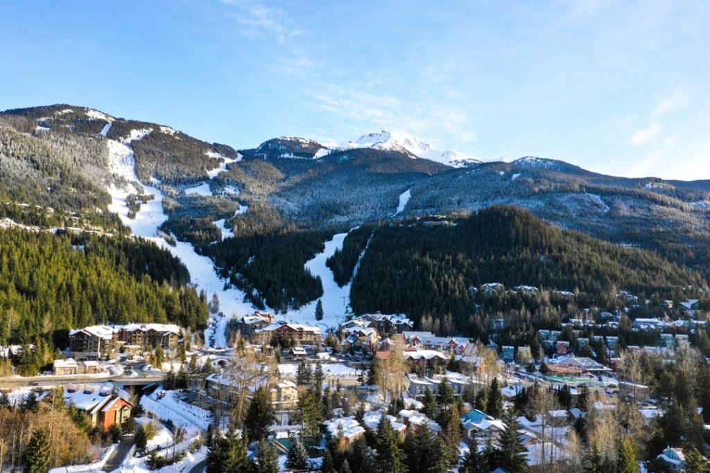 Luchtfoto van het besneeuwde dorp Whistler met kale skipistes tussen dennenbos en hoge bergtoppen op de achtergrond onder een blauwe lucht