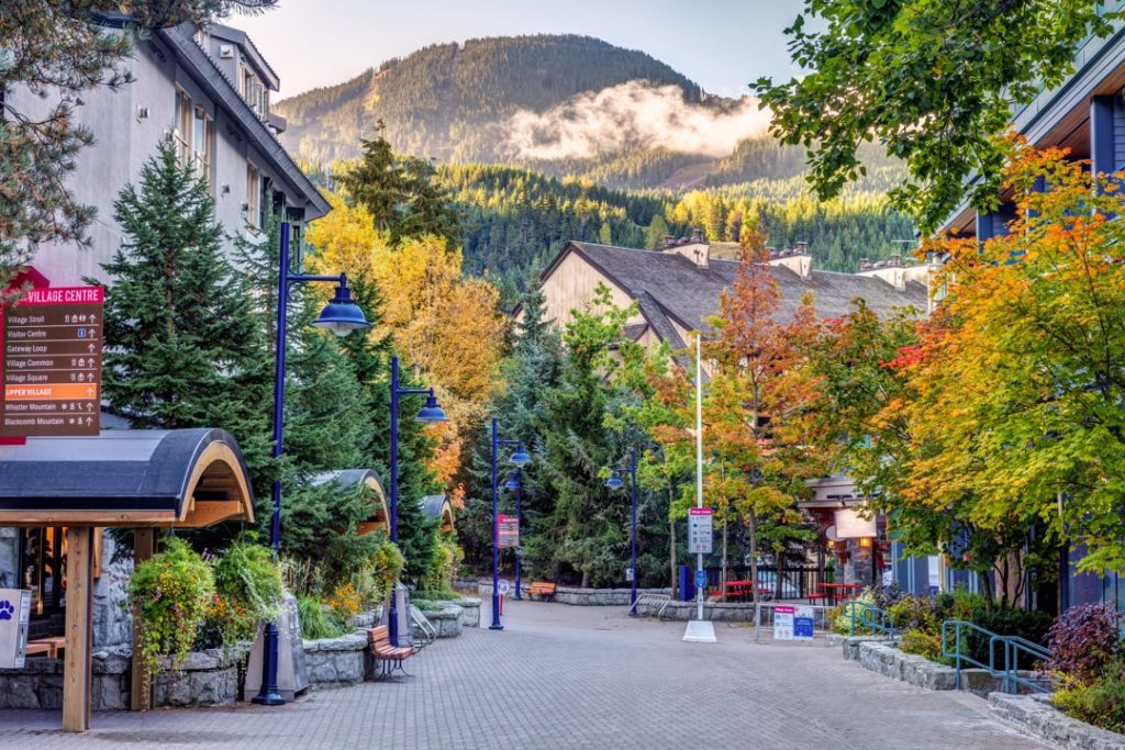 Voetgangersstraat in Whistler Village met bewegwijzeringsborden, bomen in herfstkleuren en een beboste berg die oplicht in de ochtendzon