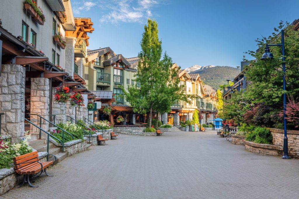 Lege bestrate voetgangersstraat in Whistler Village met natuurstenen gebouwen, hangende bloembakken, bankjes en een besneeuwde bergtop in de verte