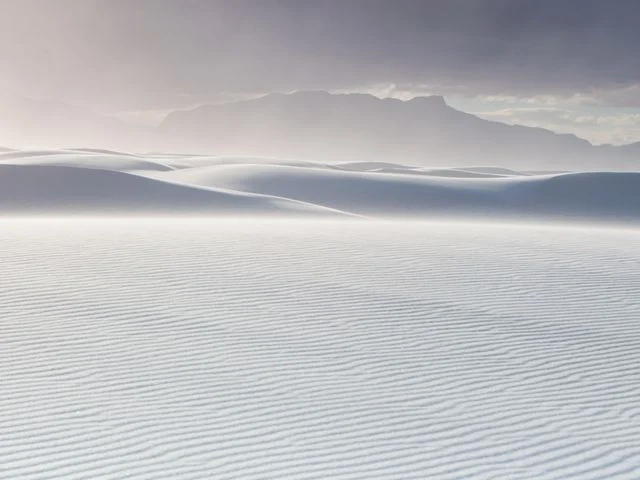 Witte gipsduinen van White Sands National Park met fijne golfpatronen in het zand en donkere bergen achter een sluier van opwaaiend zand