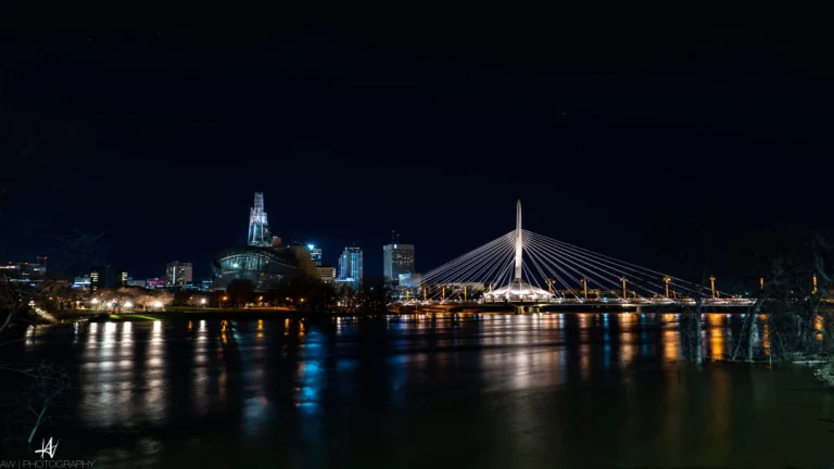 Nachtelijke skyline van Winnipeg met verlichte gebouwen, een museum en een kabelbrug die weerspiegelen in de rivier