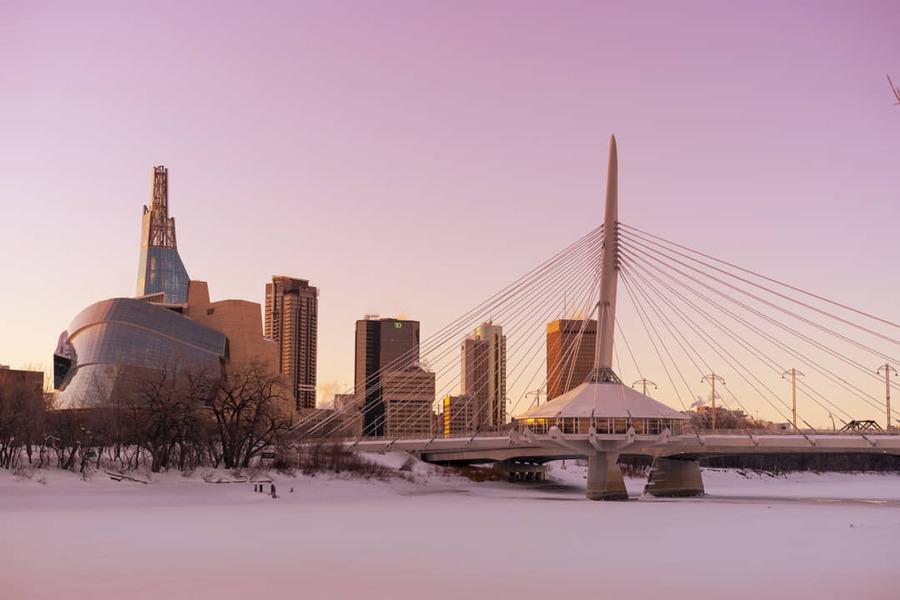Winnipeg skyline bij zonsondergang met het Canadian Museum for Human Rights, een kabelbrug en een bevroren rivier met sneeuw