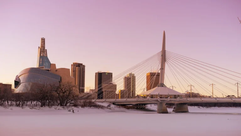 Winnipeg skyline met het mensenrechtenmuseum en een kabelbrug boven een bevroren rivier bij een roze avondlucht