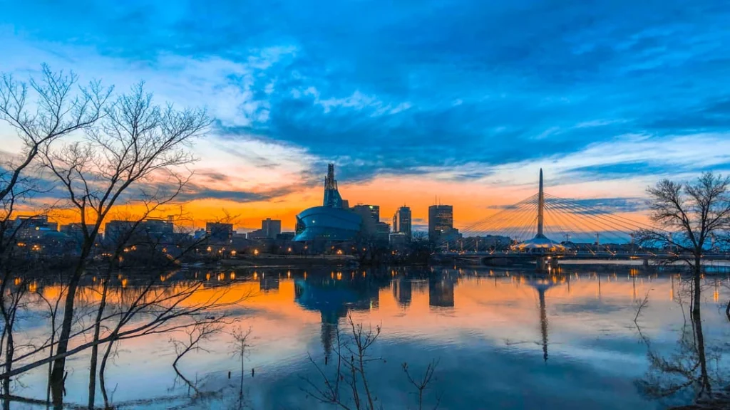 Kleurrijke ochtendlucht boven de skyline van Winnipeg met het mensenrechtenmuseum, een kabelbrug en kale bomen bij de rivier