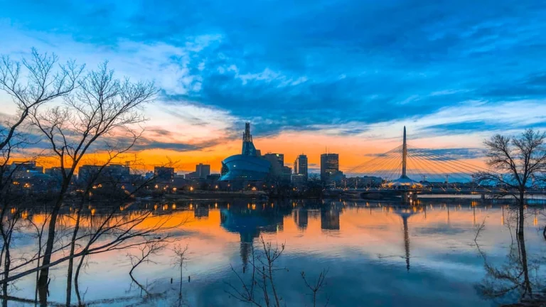 Kleurrijke ochtendlucht boven de skyline van Winnipeg met het mensenrechtenmuseum, een kabelbrug en kale bomen bij de rivier