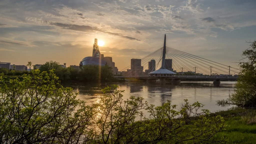 Gouden zonsondergang boven de Red River met de skyline van Winnipeg en een kabelbrug achter groene struiken
