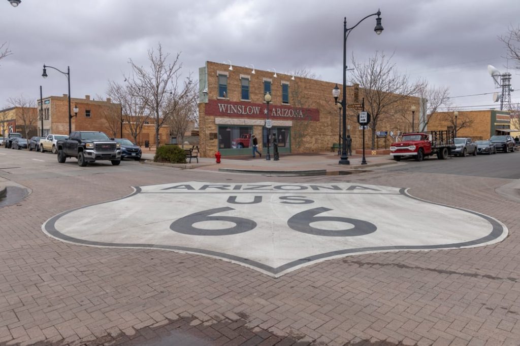 Groot wegschild van Route 66 geschilderd op straat, met op de achtergrond het gebouw met de tekst Winslow Arizona