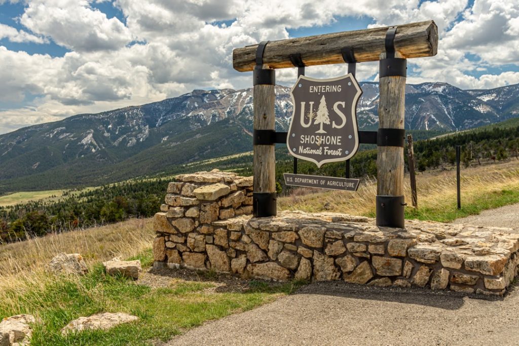 Houten bord met stenen voet dat de ingang van het Shoshone National Forest markeert, met een uitgestrekt berglandschap op de achtergrond