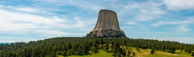 De kolomvormige rotstoren van Devils Tower rijst op boven een glooiende heuvel vol dennenbomen onder een blauwe lucht met sluierwolken