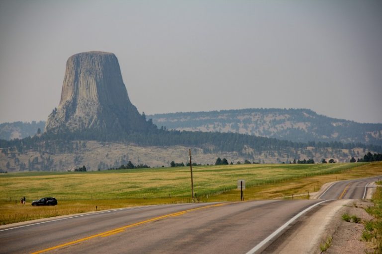 Devils Tower rijst op boven glooiende graslanden en bomen, gezien vanaf een lege tweebaansweg door het platteland