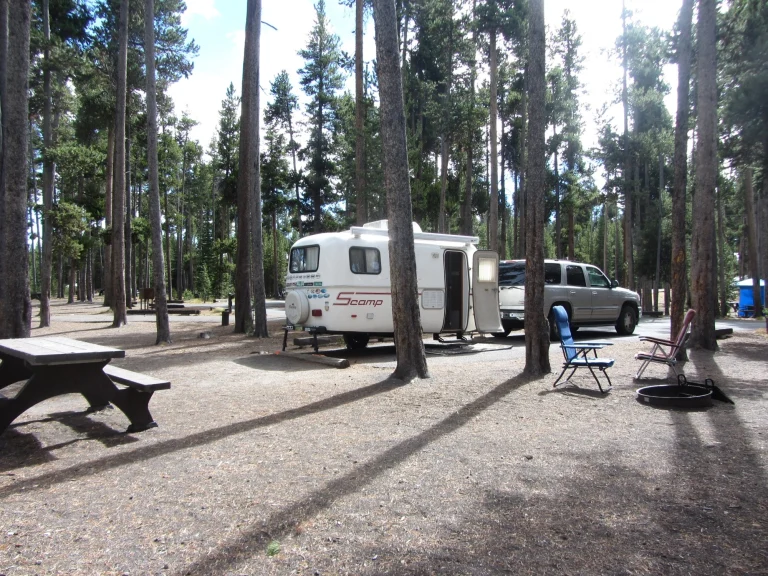Kleine witte camper gekoppeld aan een suv staat op een kampeerplek tussen hoge dennenbomen met kampeerstoelen en een vuurplaats ernaast