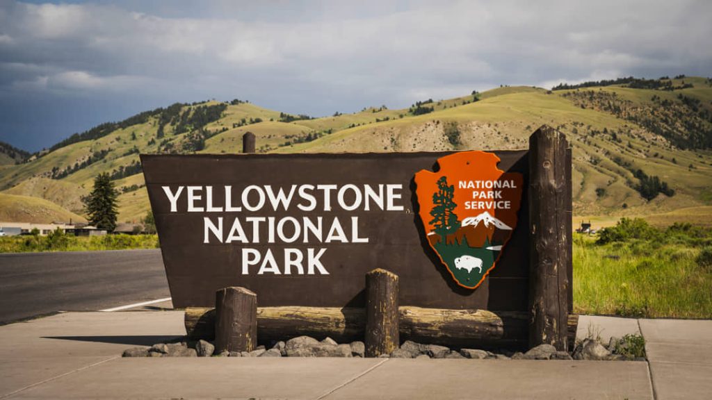 Groot bruin bord met het opschrift Yellowstone National Park en het logo van de National Park Service, met glooiende heuvels op de achtergrond