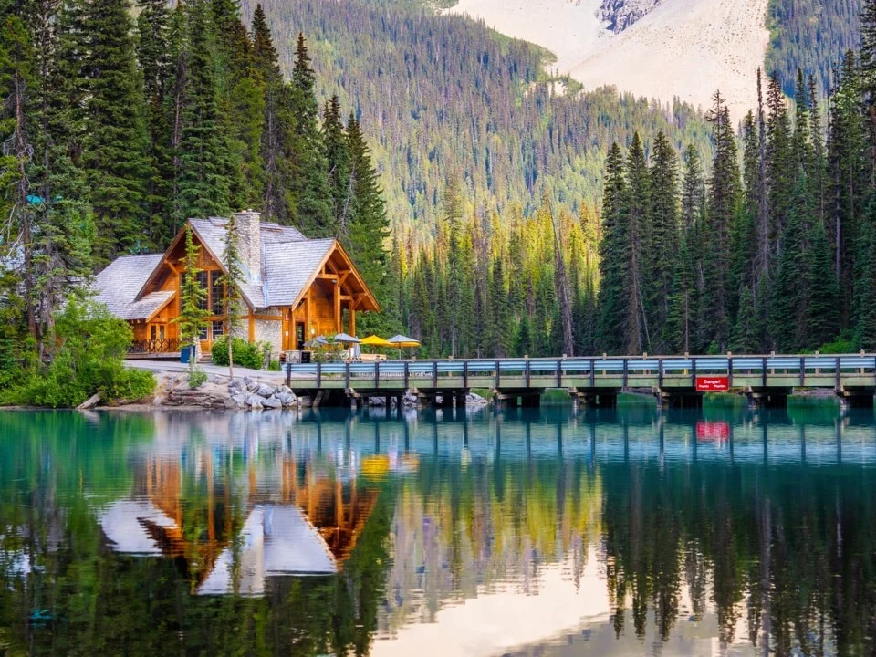 Een houten lodge met terras en een brug over turkoois water, omringd door dennenbos en besneeuwde bergen in Yoho National Park
