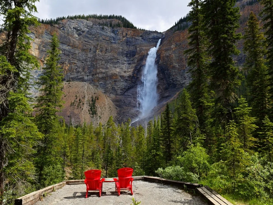 Twee rode houten stoelen staan op een uitkijkplatform met zicht op de hoge Takakkaw Falls omringd door dennenbos in Yoho National Park
