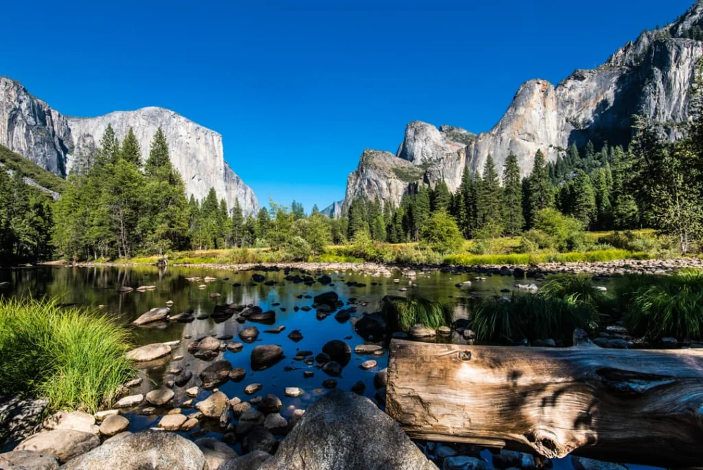 Grijze granieten rotswanden van Yosemite Valley weerspiegelen in een rustige rivier met rotsen en bomen op de voorgrond