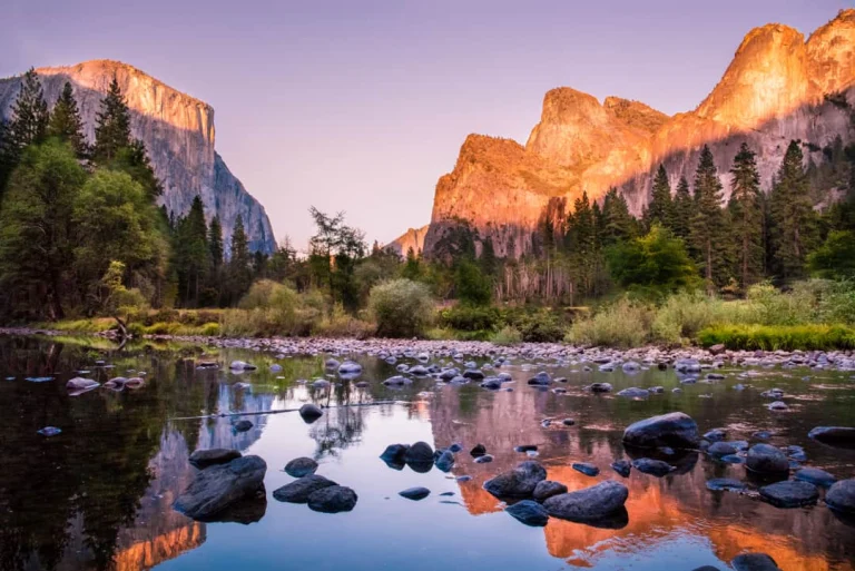 Ondergaande zon werpt een gouden gloed over de rotswanden van Yosemite National Park, met een rustige rivier vol keien op de voorgrond