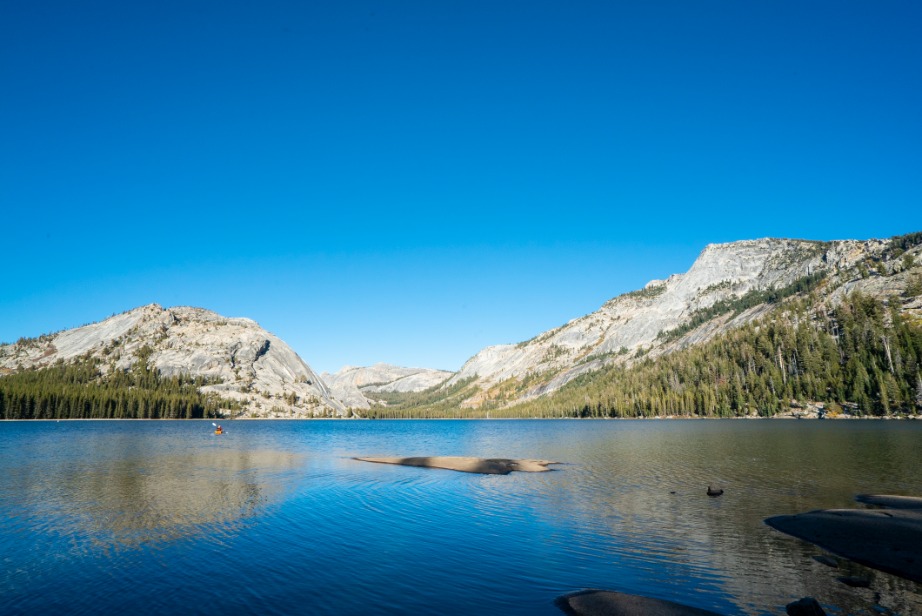 Helder blauw bergmeer met een persoon die erin staat, omringd door kale granieten bergtoppen en dennenbos in Yosemite National Park