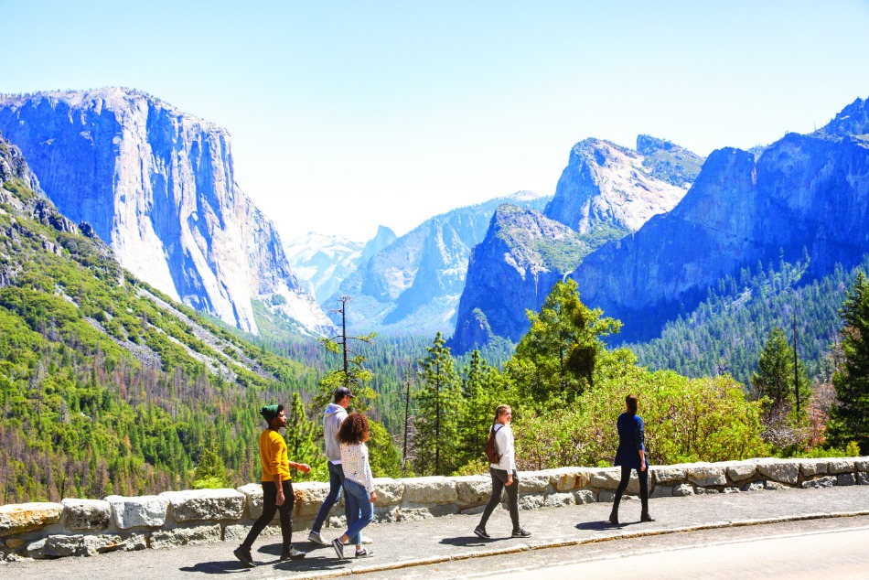 Groepje wandelaars loopt langs een stenen muurtje bij het uitkijkpunt Tunnel View met zicht op de bergen van Yosemite Valley