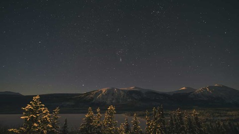Heldere sterrenhemel boven besneeuwde bergtoppen en een meer met dennenbomen op de voorgrond in Yukon
