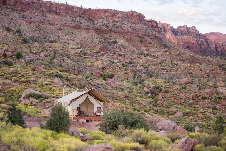 Een witte safaritent met houten terras staat tussen rotsen en struiken onder de roodgekleurde bergwand van Zion National Park