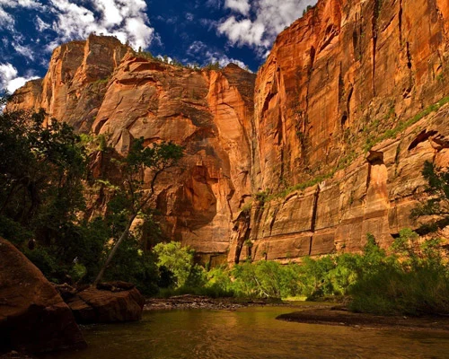 Hoge rode en oranje rotswanden van Zion National Park torenen boven een kalme rivier omzoomd door groene begroeiing en rotsblokken